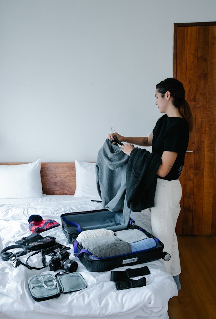 A Woman in Black T-shirt Packing Clothes Beside a Bed with Suitcase